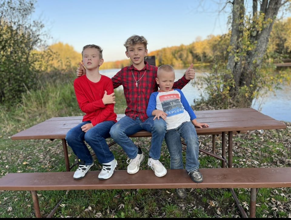 Three brothers are sitting on a picnic bench in front of a lake. 